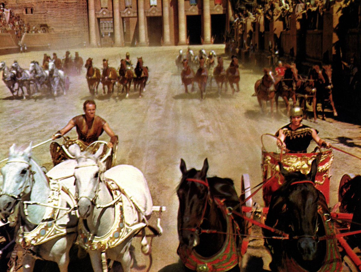 A bustling ancient Roman arena filled with chariots and horses, with spectators lining the stands and a central dirt track where multiple chariots race past.