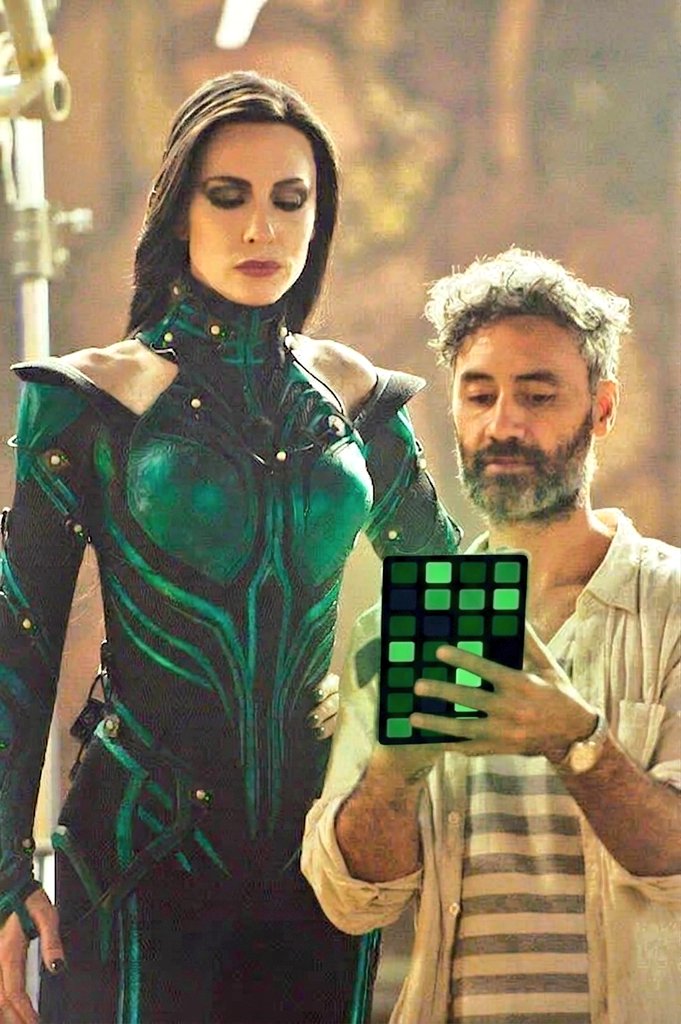 A woman in a detailed green and black armored costume stands beside a man with a beard and gray hair, who is holding a tablet displaying a grid of green squares. The man appears to be reviewing or adjusting the color palette on the device.
