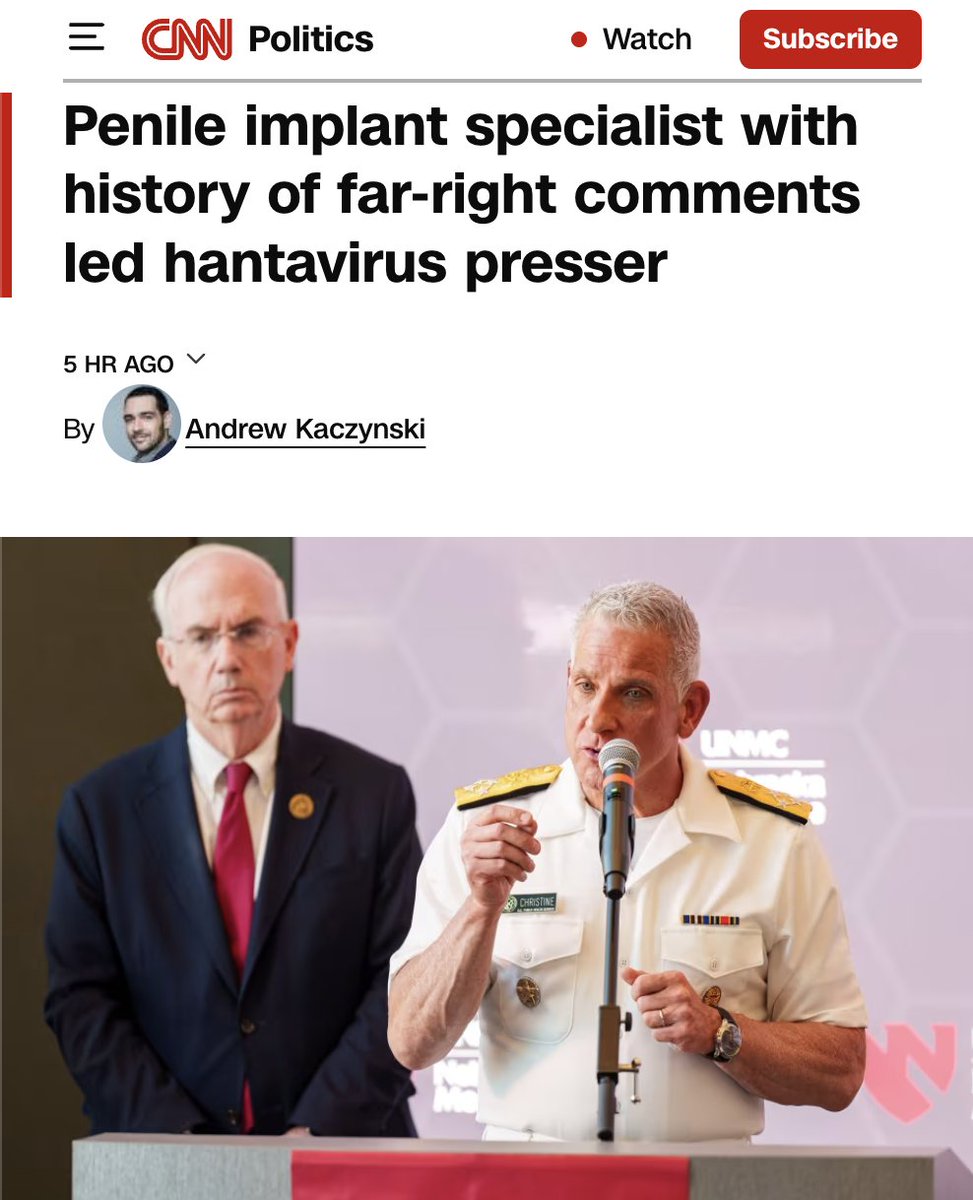 A man in a white military uniform with medals and a name tag is speaking into a microphone at a podium, while another man in a dark suit and red tie stands beside him with a serious expression. The background features a hexagonal pattern and partial logos, including "UNMC".