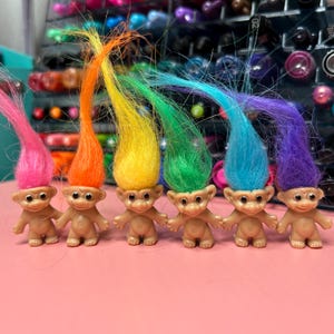 A row of five small troll figurines with vibrant, rainbow-colored hair—pink, orange, yellow, green, and blue—stands on a pink surface, with a blurred background of colorful objects.