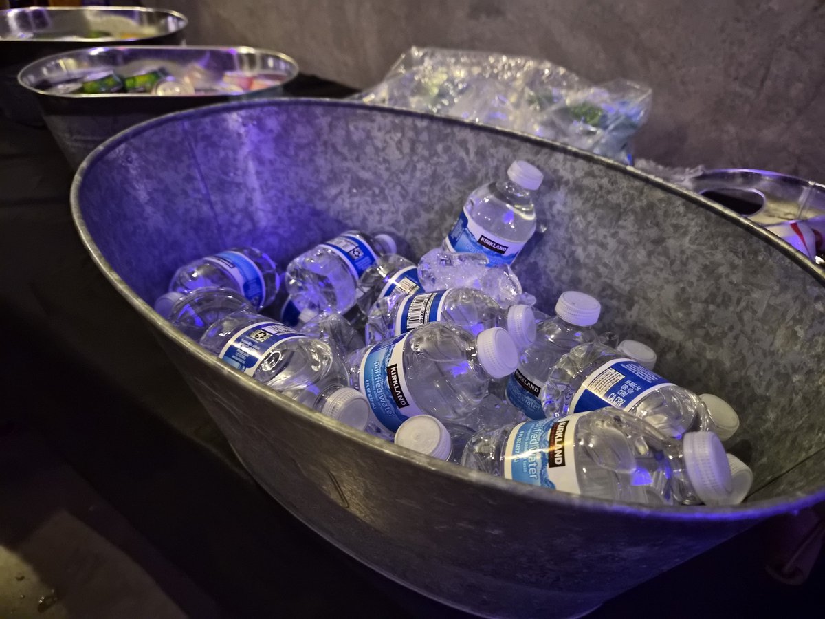 A large galvanized metal tub filled with ice and numerous Kirkland brand water bottles, illuminated by a cool blue light, sits on a dark surface with other similar containers in the background.