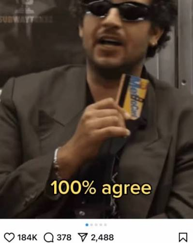 A man with curly hair and sunglasses, wearing a dark suit, holds up a small object resembling a credit card. Yellow text overlaid on the image reads "100% agree".