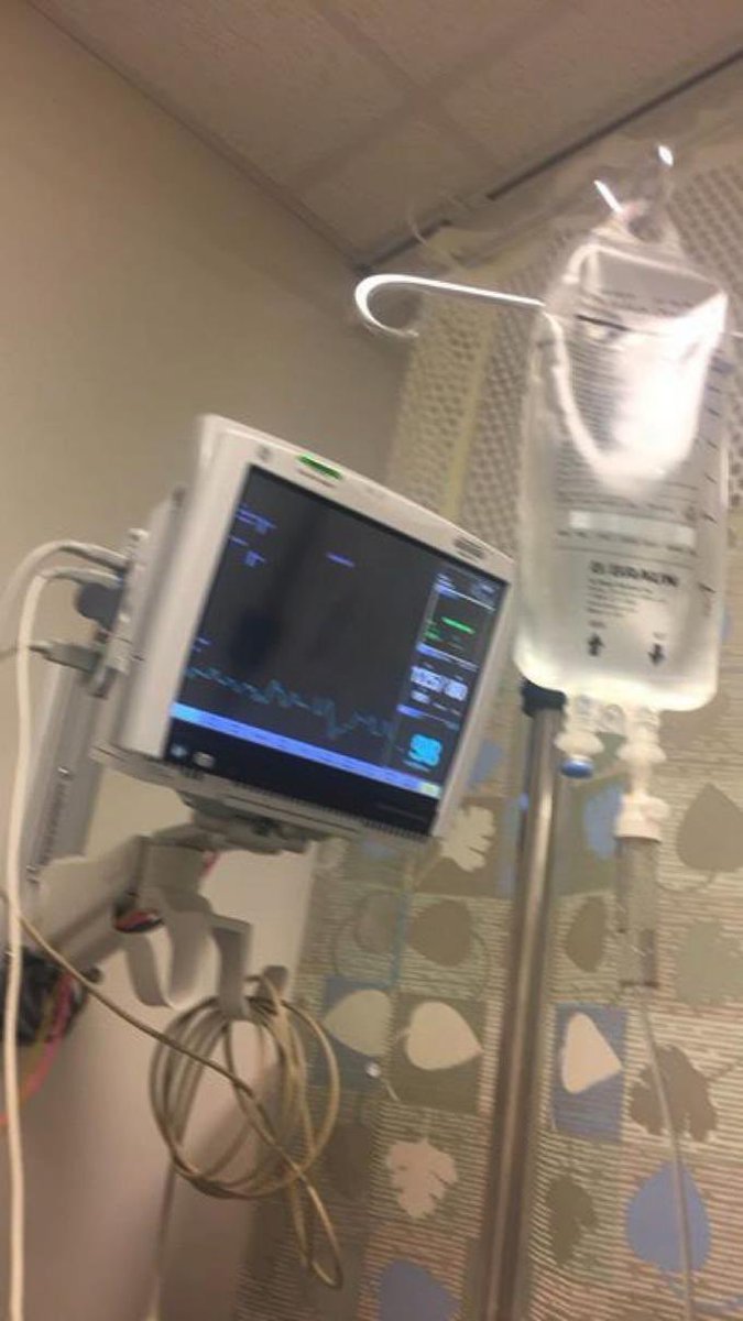 A medical monitor displaying vital signs with a blue waveform and numerical readouts, positioned next to an IV bag hanging from a stand in a hospital room with patterned curtains.