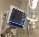 A medical monitor displaying vital signs is mounted on a stand in a hospital room, with an IV bag hanging nearby.