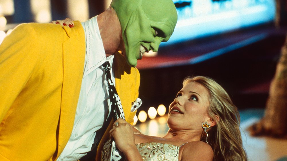 A man with a bright green face and a wide grin wearing a yellow suit leans over a blonde woman in a sparkling dress. The woman looks up at him while holding his black and white polka dot tie.