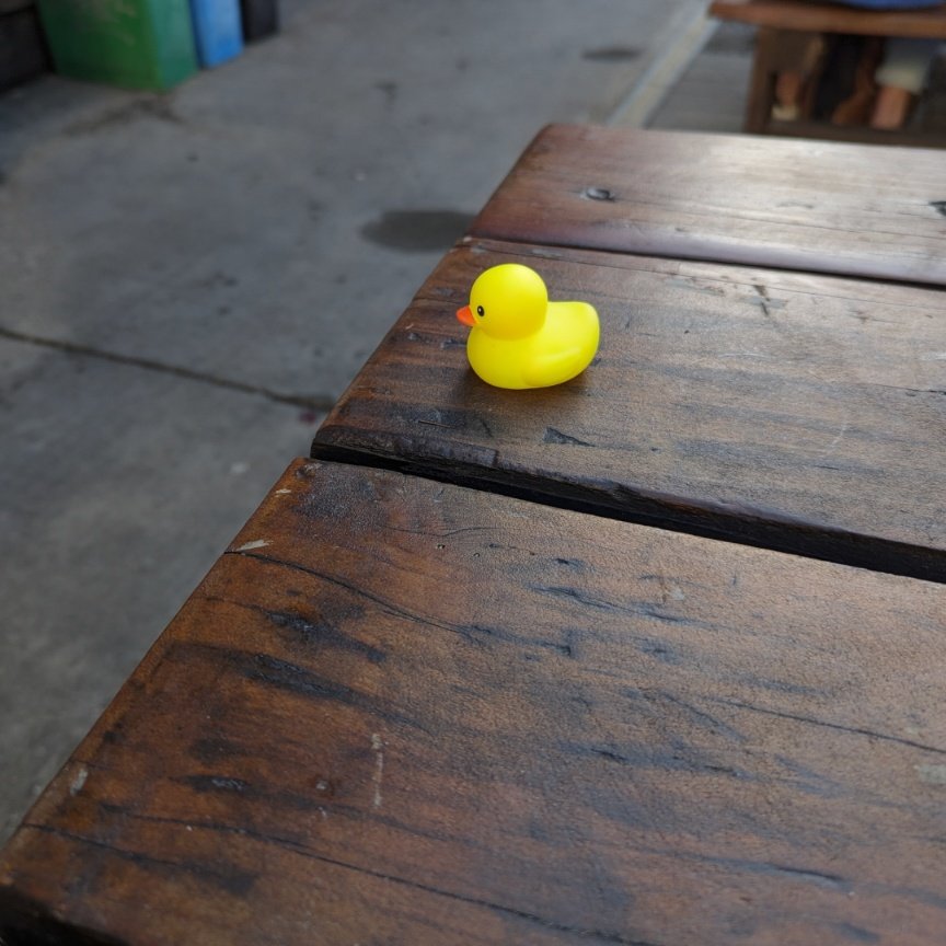 A small yellow rubber duck with an orange beak sits on the edge of a dark wooden picnic table. The background shows a blurred concrete ground and the legs of another wooden table.