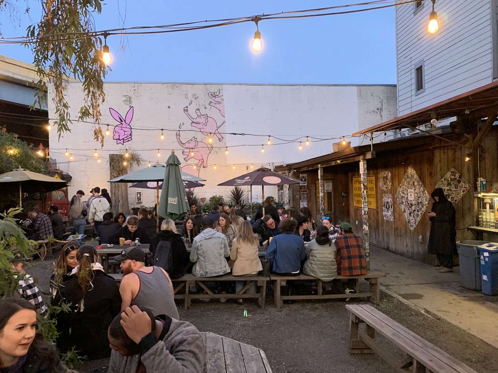 A crowded outdoor patio at dusk with people sitting at long wooden picnic tables under string lights and umbrellas. A white wall in the background features pink graffiti of a bunny head and elephants.