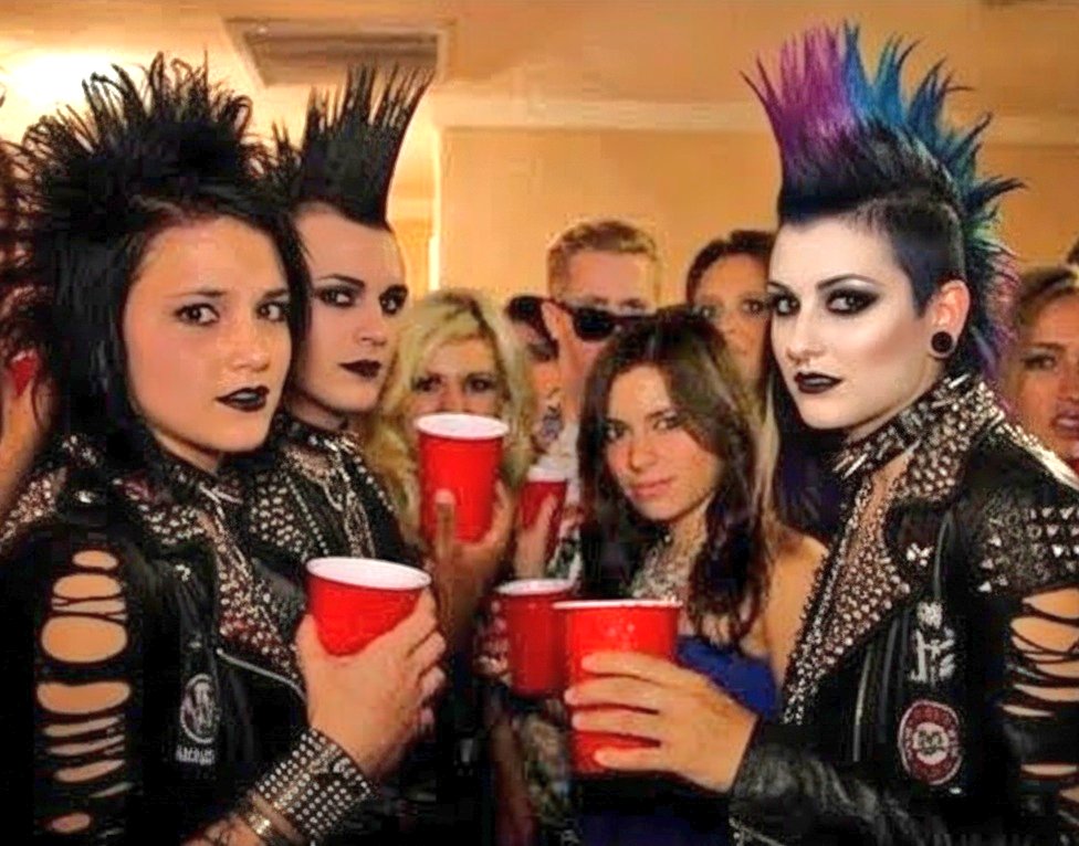 A group of people at a party, featuring three individuals in the foreground with tall punk hairstyles, dark makeup, and studded leather jackets. They are holding red plastic cups and posing for the camera alongside other partygoers in the background.