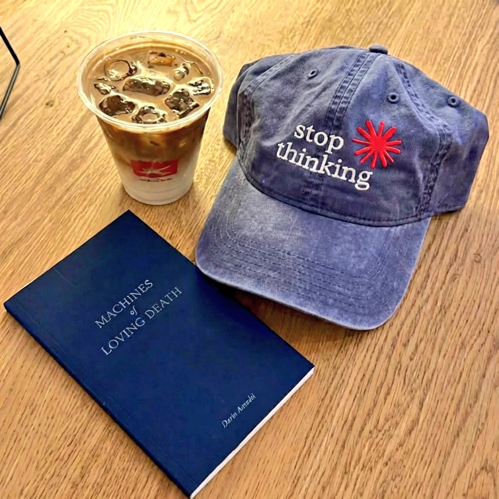 On a wooden table, a blue cap with "Stop thinking" and a red star logo sits next to a blue book titled "Machines of Loving Death" by Darni Amadei and an iced coffee with ice cubes in a clear plastic cup.