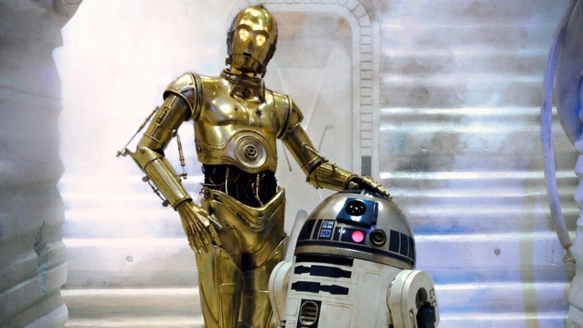 C-3PO, a tall golden droid, stands beside R2-D2, a smaller white and blue droid, on a staircase in a brightly lit, futuristic setting.